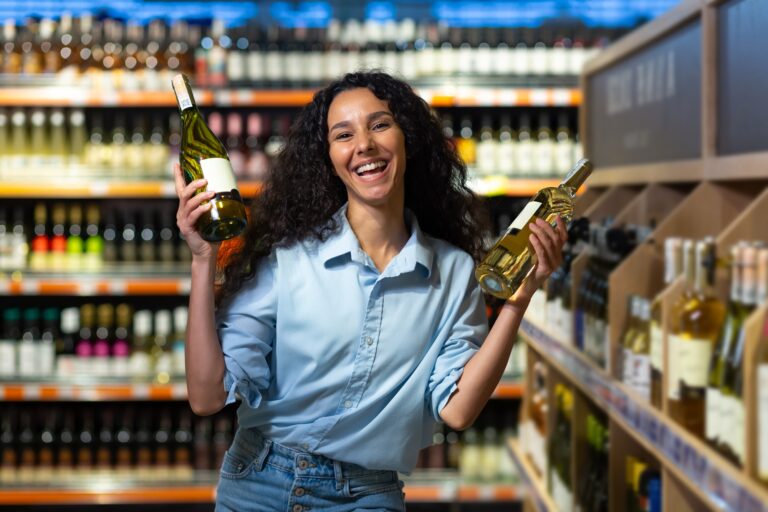 Joyful woman choosing wine bottles from liquor store shelves smiling